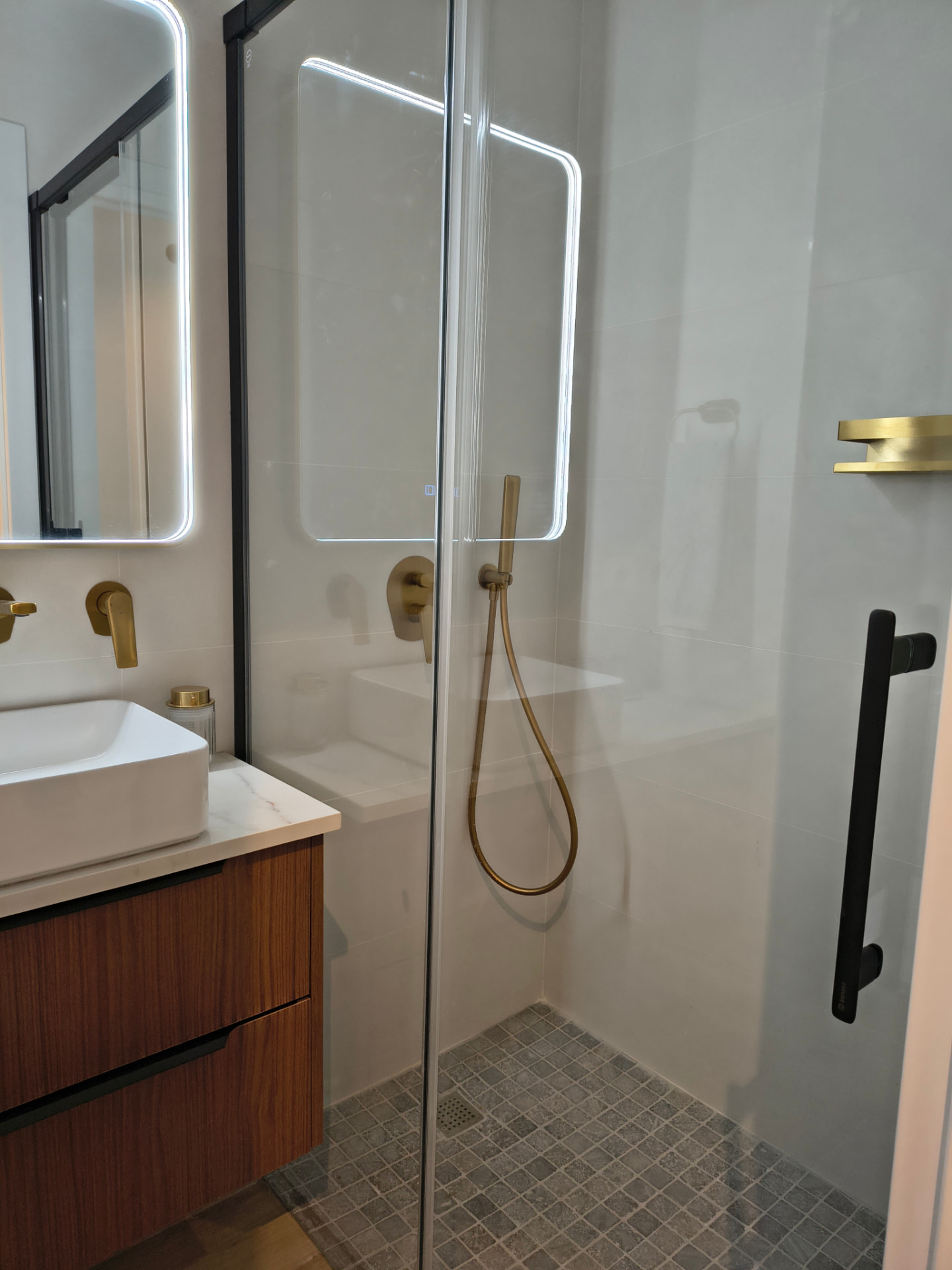 Baño con ducha de obra, mampara negra, grifería dorada y mueble de nogal